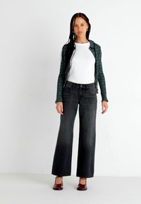 Jeans anchos negros combinados con una camiseta blanca y una chaqueta de cuadros verdes. El conjunto incluye zapatos burdeos y tiene el cabello largo y liso.