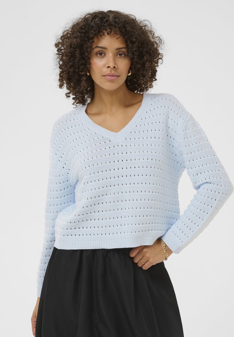 Femme aux cheveux bouclés portant un pull léger en maille bleu clair ajouré et une jupe noire, posant avec la main sur la hanche devant un fond uni.