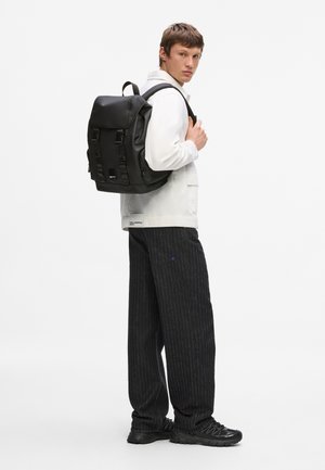 Tânăr îmbrăcat într-o jachetă albă și pantaloni cu dungi subțiri, purtând un rucsac negru mare peste un umăr, stând pe un fundal alb.