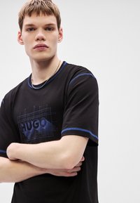Camiseta preta com costura de realce azul e estampa gráfica com a palavra "HUGO." Mangas curtas e decote redondo. Textura de tecido macio.