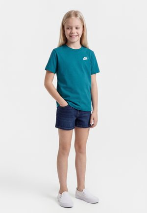 Uśmiechnięta dziewczyna z długimi blond włosami ma na sobie turkusowy t-shirt marki Nike, ciemne jeansowe szorty i białe wsuwane buty, stoi z jedną ręką w kieszeni.