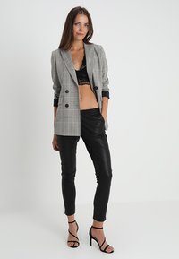 Blazer doppiopetto a scacchi grigi sopra un crop top in pizzo nero, abbinato a pantaloni aderenti in ecopelle nera e sandali neri con cinturini.