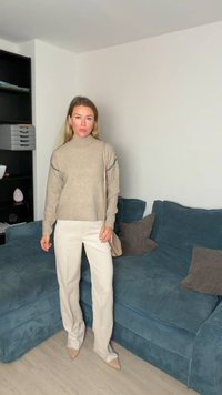 Camisola bege em malha com gola alta e detalhe em ponto contrastante, combinada com calças brancas de perna reta e saltos cor de pele. Sofá azul ao fundo.