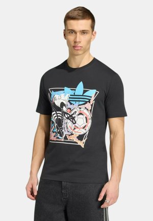 Giovane uomo con t-shirt grafica nera raffigurante uno scarpino da calcio, una palla e il logo Adidas blu, in piedi davanti a uno sfondo semplice.