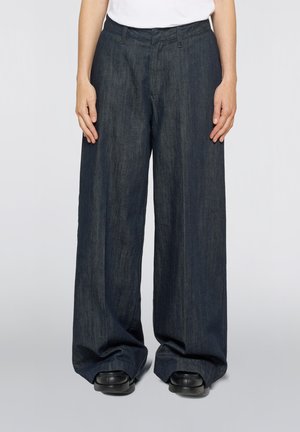 Person iført mørke vide denimbukser og sorte sko med hvid top, stående foran en ensfarvet lys baggrund.