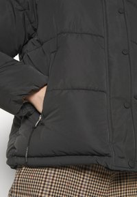 Μαύρο μπουφάν με επένδυση, με ραφές σε πλέγμα, που διαθέτει κουμπιά με κλιπ και τσέπη με φερμουάρ. Φορεμένο πάνω από παντελόνι με καρό σχέδιο.