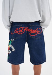 Shorts de mezclilla con un lavados azul, con el lettering rojo de "Ed Hardy" en la parte trasera y un diseño gráfico colorido en la pierna izquierda.