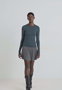 Langärmliges türkisfarbenes Oberteil aus glattem Stoff, kombiniert mit einem grauen, plissierten Rock. Das Model trägt kniehohe schwarze Stiefel und steht vor einem neutralen Hintergrund.