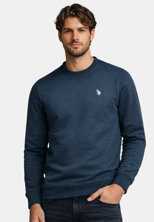 Man with wavy brown hair wearing a navy blue crewneck sweatshirt with a small light blue embroidered logo on the chest.