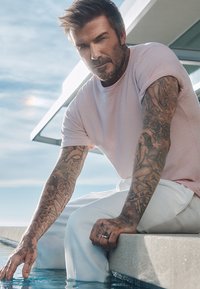 Ljust rosa kortärmad skjorta och vita byxor bärs av en person som sitter vid kanten av en pool, med detaljerade tatueringar synliga på armarna.