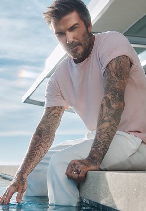 Light pink short-sleeve shirt and white pants worn by a person sitting at an edge of a pool, with detailed tattoos visible on arms.
