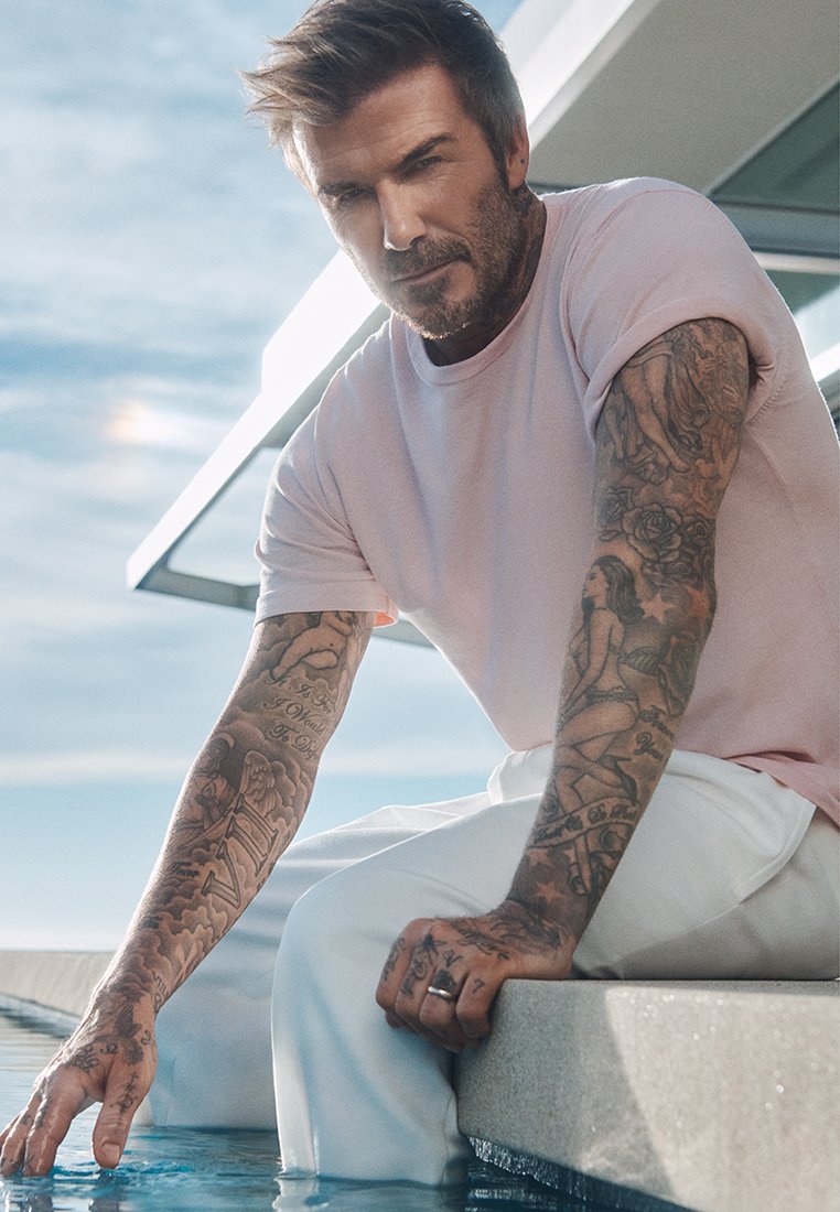 Ljust rosa kortärmad skjorta och vita byxor bärs av en person som sitter vid kanten av en pool, med detaljerade tatueringar synliga på armarna.