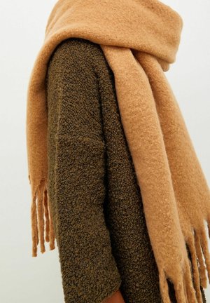 Personne portant un manteau marron texturé avec une grande écharpe beige douce enroulée autour du cou, avec des franges pendantes sur un côté.