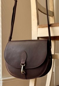 Sac en cuir brun à bandoulière avec une forme arrondie, une sangle unique, une fermeture à rabat et des accessoires en métal doré. Texture lisse, design minimaliste.