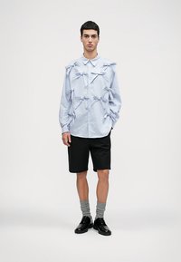 Ανοιχτό μπλε ριγέ πουκάμισο με φιόγκους, μαύρες βερμούδες, γκρι κάλτσες και μαύρα παπούτσια. Το πουκάμισο διαθέτει γιακά με κουμπιά και μακριά μανίκια.