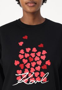 Sweatshirt preta com padrões de corações vermelhos e rosas, com bordado texturizado e uma inscrição ousada em branco a dizer "Karl" na parte inferior.