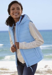 Helles, blau gepolstertes Weste mit Kapuze, Reißverschluss vorne und zwei seitlichen Taschen, über einem cremefarbenen Langarmoberteil getragen. Kombiniert mit einer dunkelblauen Jeans.