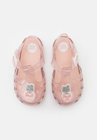 Sandalias translúcidas rosas con brillantina, con una correa y un charm en forma de oso que dice 'querido' en un corazón. Diseño moldeado y abierto en los dedos.