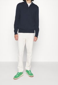 Pull-over bleu marine avec col à demi-fermeture éclair, associé à un pantalon couleur crème et des baskets vertes avec des lacets blancs et une semelle en gomme.