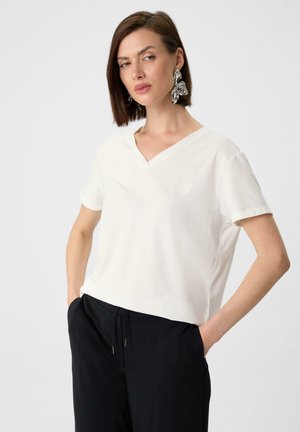 T-shirt blanc à col en V et manches courtes avec un logo discret sur la poitrine, associé à un pantalon noir. Le tissu est lisse et léger.