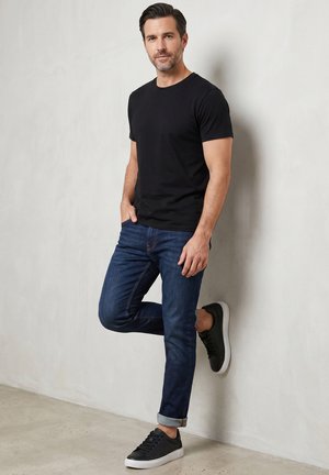 Man met een zwart T-shirt, donkerblauwe opgerolde spijkerbroek en zwarte sneakers, leunend tegen een witte muur met één been opgetild.