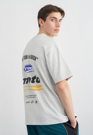 Młody mężczyzna z krótkimi brązowymi włosami, ubrany w szary t-shirt z nadrukiem i turkusowe spodnie, spoglądający przez ramię na białym tle.