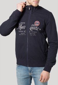 Marineblå zip-up jacka i blandmaterial av bomull, med vitt tryckt text och en rund logotypapplikation. Slät textur, figurnära design och långa ärmar.
