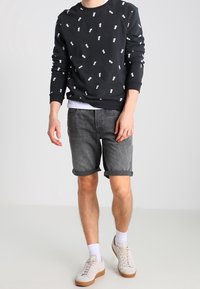 Czarny sweatshirt z białym nadrukiem ananasa, szare dżinsowe szorty z podwiniętymi nogawkami oraz jasne tenisówki z beżową podeszwą na neutralnym tle.
