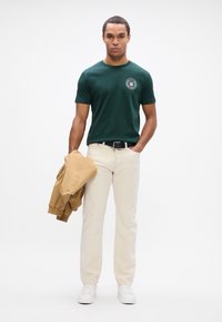 Tumedast roheline lühikeste varrukatega t-särk ümmarguse logoga, beežid chino püksid ja khaki bomber-jakk vasakus käes, valged spordijalatsid jalas.