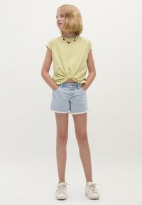 Hellgelbes, kurzärmliges Oberteil mit Frontknoten. Blaue Jeansshorts mit fransigem Saum und drei Knöpfen. Weiße Sneaker.