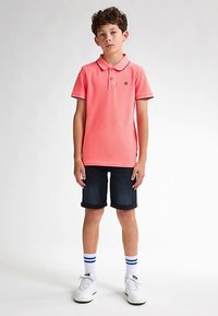 Růžové polo tričko s límečkem a logem, kombinované s tmavými džínovými šortkami. Bílá teniska se zelenými akcenty a proužkované ponožky doplňují vzhled.