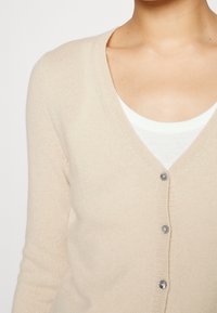 Beige Cardigan mit V-Ausschnitt, fünf Knöpfen und langen Ärmeln; weiche Textur, leichtes Material, mit subtilen Rippendetails.