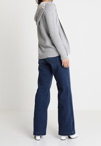 Kvinna som bär en grå huva-sweatshirt, mörkblå vida jeans och vita klackskor, stående mot en helt vit bakgrund.