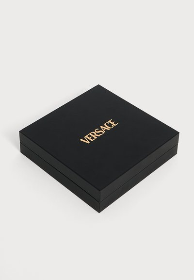 Boîte carrée noire fermée avec le logo Versace doré centré sur le dessus, sur un fond clair et uni.