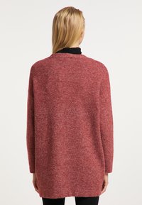 Pulover lung, lejer, de culoare roșie, realizat dintr-un material tricotat texturat, cu un guler rotund și umeri căzuți, purtat peste pantaloni negri.