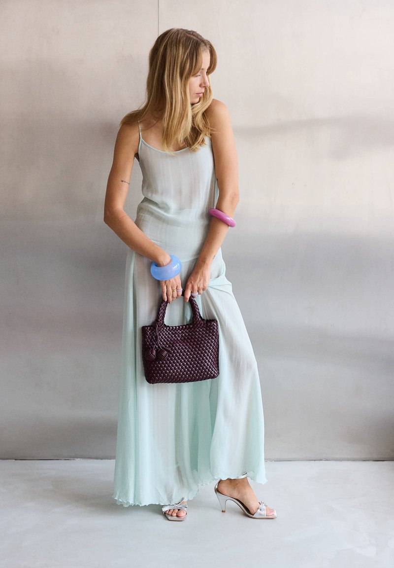 Frau in einem blassgrünen ärmellosen Kleid, die eine strukturierte dunkelviolette Handtasche hält, trägt blaue und pinke Armreifen und weiße Sandalen mit hohen Absätzen.