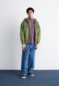 Olive green zip-up hoodie with front pockets, layered over a lavender t-shirt. Loose, light blue denim pants and green slip-on shoes.