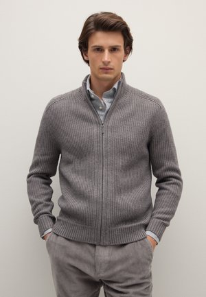 Junger Mann steht mit in den Taschen vergrabenen Händen, trägt einen grauen, gerippten Reißverschluss-Pullover über einem Hemd mit Knöpfen und graue Cordhosen.