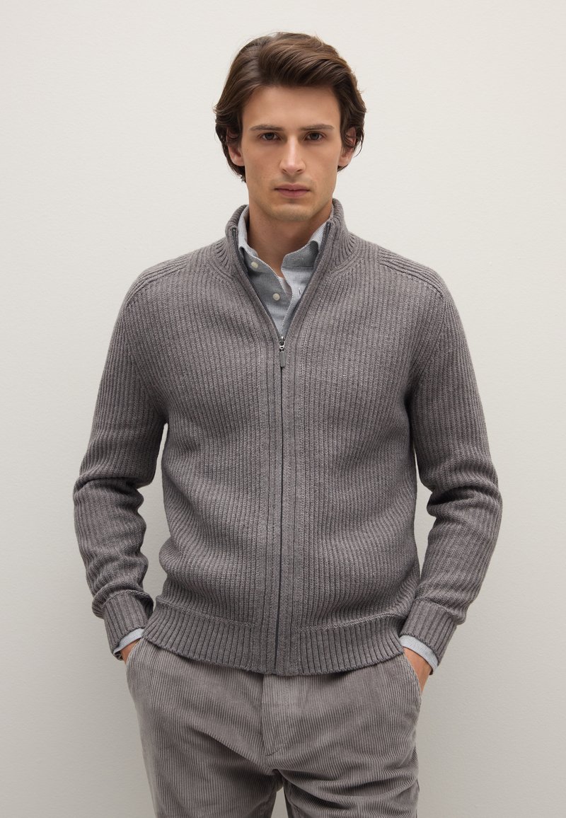Jeune homme debout les mains dans les poches, portant un pull gris côtelé zippé sur une chemise boutonnée et un pantalon en velours côtelé gris.