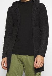 Cardigan lungo nero lavorato a maglia con cappuccio, caratterizzato da un motivo a trama. Indossato sopra una maglietta nera e pantaloni verde oliva.