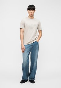 Ανοιχτό γκρι κοντομάνικο μπλουζάκι με ανάγλυφη λεπτομέρεια λογότυπου, σε συνδυασμό με χαλαρά, μεσαίου μπλε τζιν και μαύρα παπούτσια.