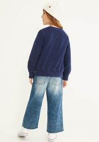 Felpa blu navy con polsini e orlo a coste, abbinata a jeans in denim sbiaditi a gamba larga e sneaker bianche. Cappello di maglia bianco in testa.