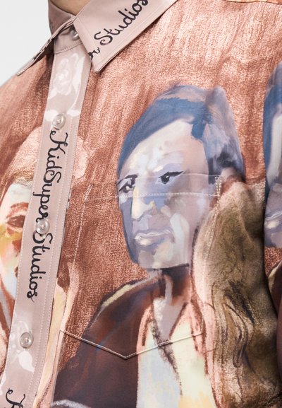 Πουκάμισο με κοντά μανίκια και κουμπιά σε πολύχρωμο σχέδιο με πορτρέτα, με γυαλιστερή υφή. Ετικέτα "KidSuper Studios" στον γιακά.