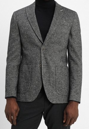 Homme portant un blazer gris en laine ajusté sur un col roulé noir et un pantalon noir, debout devant un fond uni.