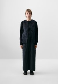 Zwarte vest met meerdere zakken, over een zwarte lange mouwen shirt en brede zwarte broek, allemaal gemaakt van een gladde, matte stof.