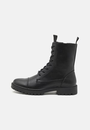 Botte de combat en cuir noir avec laçage à l’avant, languettes en haut et semelle épaisse texturée, vue de côté sur fond blanc.