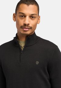 Sort strikket zip-up sweater med høj krave og subtilt logo. Teksturdet stof, tætsiddende design, og lagdelt over en grøn skjorte.