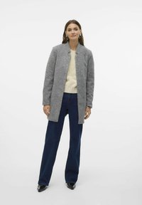 Grau wollener Mantel mit geradem Schnitt, Stehkragen und ohne sichtbare Verschlüsse, kombiniert mit einem cremefarbenen Kabelstrickpullover und dunkelblauen Jeans.