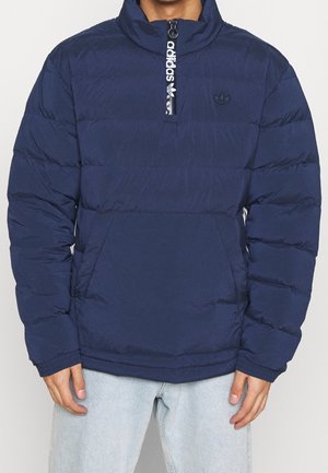 Doudoune bleu marine Adidas avec demi-fermeture éclair et poche kangourou, portée avec un jean bleu clair, mains détendues sur les côtés.