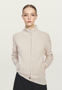 Maglione a coste beige chiaro con colletto alto, zip frontale completa e maniche lunghe, caratterizzato da una texture morbida e una silhouette aderente.